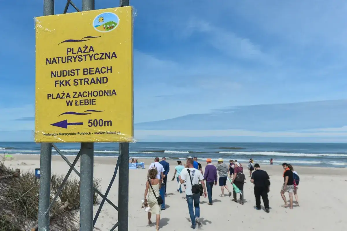 Ż&oacute;łty znak informuje: plaża naturyistyczna, nudist beach, FKK Strand. Ludzie idą w stronę morza.