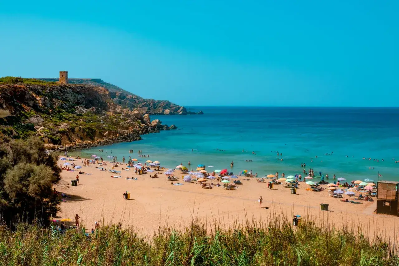 Malta plaże piaszczyste z turkusową wodą i ludźmi odpoczywającymi pod parasolami. Na wzgórzu widać starą wieżę.