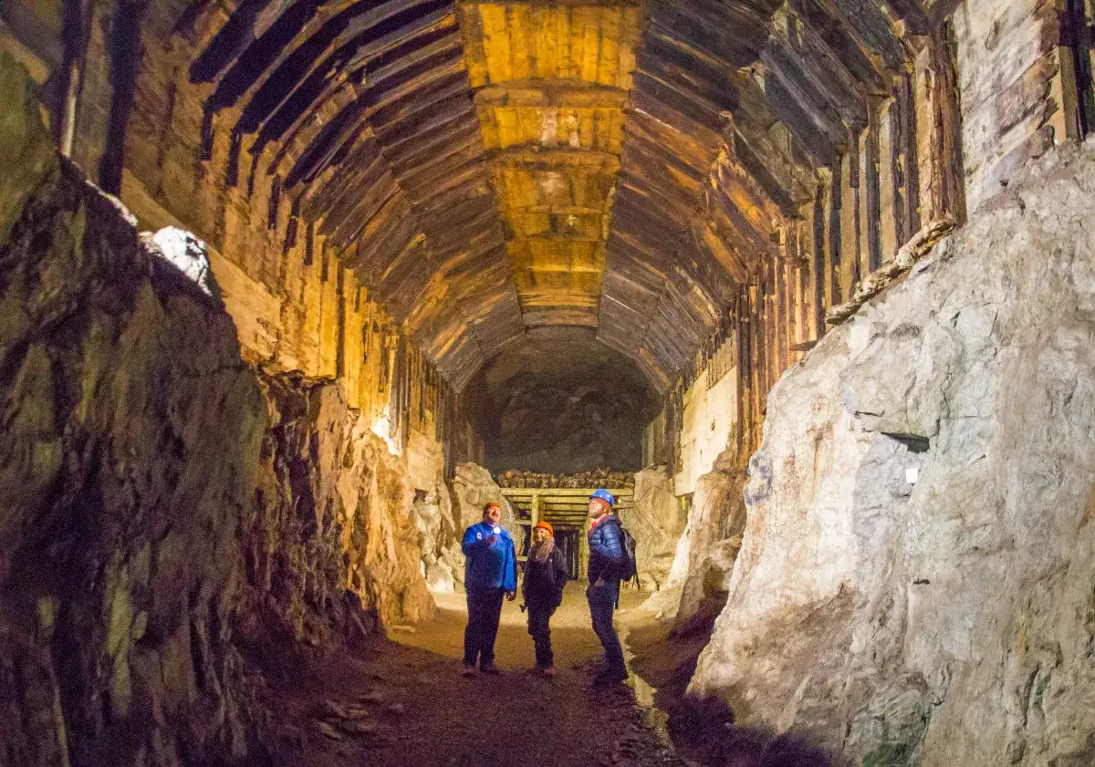 Trzech turyst&oacute;w w kaskach oświetla latarkami mroczny tunel kopalni. Czy to os&oacute;wka czy włodarz, trudno stwierdzić w tym labiryncie.