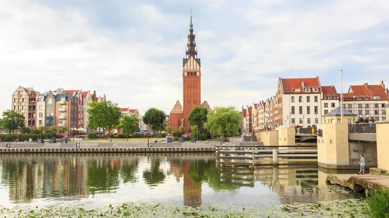 Elbląg atrakcje: nadbrzeże z kolorowymi kamienicami i wieżą kościoła św. Mikołaja, odbijające się w spokojnej wodzie.