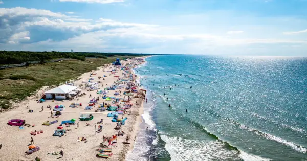 Tłum ludzi na plaży w Darłowie, korzystających z uroków Bałtyku. Wiele kolorowych parawanów i namiotów, a w wodzie kąpiący się. To typowe atrakcje Darłowa latem.