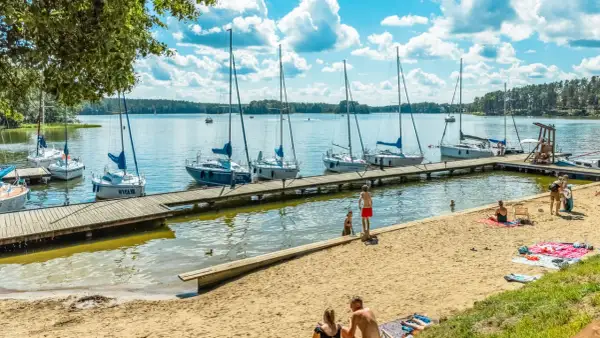 Słoneczny dzień na mazurskich jeziorach. Cumujące jachty, piaszczysta plaża i ludzie odpoczywający nad wodą.