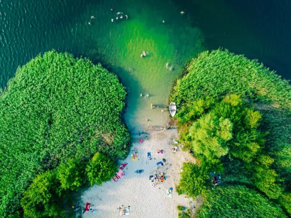Letnie, magiczne jeziora w Polsce. Ludzie kąpią się w zielonej wodzie, odpoczywają na piaszczystej plaży otoczonej trzcinami i drzewami.