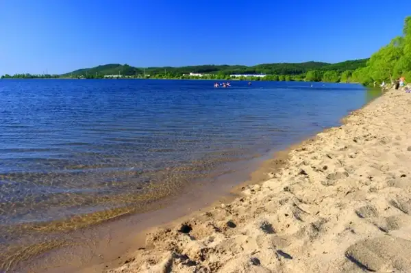 Słoneczny dzień nad zalewem Sulejowskim. Piaszczysta plaża, błękitna woda i zielone wzgórza w tle.