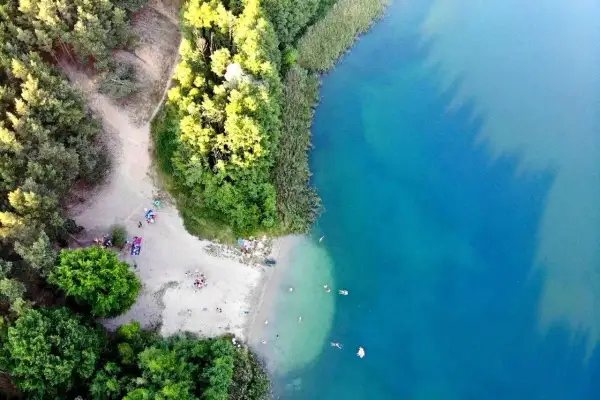 Plaża nad jeziorem tętni życiem. Ludzie odpoczywają na piasku, kąpią się w krystalicznie czystej wodzie, otoczeni zielenią drzew.
