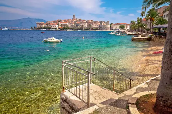 Widok na turkusowe morze i kamienistą plażę z kamiennymi schodami prowadzącymi do wody. W oddali widać urokliwe miasto Korcula, pełne zabytków i atrakcji.