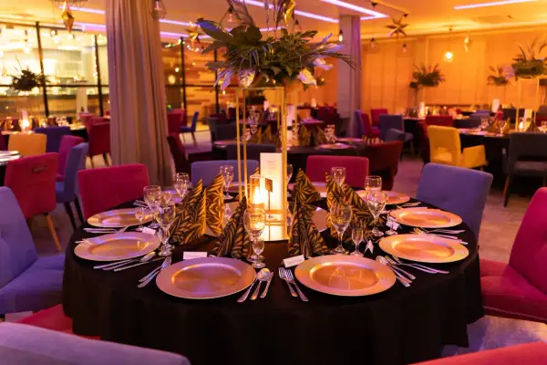 Elegancko nakryte stoły w restauracji, gotowe na przyjęcie gości. W centralnym punkcie stołu znajduje się ozdobna kompozycja z liści i świeca, tworząc ciepłą atmosferę. Kolorowe krzesła dodają wnętrzu charakteru. To idealne miejsce na wykwintne jedzeni...