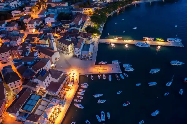 Nadmorskie miasteczko nocą, z oświetlonymi domami i portem pełnym łodzi. W oddali widać fazana plaże.