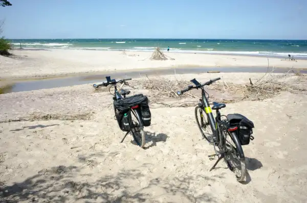 Dwa rowery z sakwami na piaszczystej plaży, gotowe na odkrywanie tras rowerowych nad morzem. W tle widać morze i niebo.
